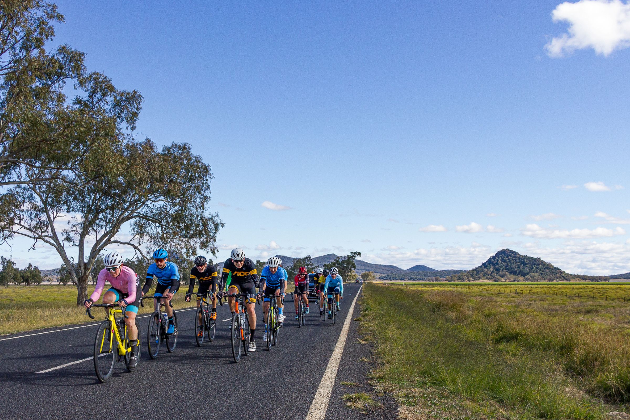 A group of 10 or so road cyclists riding on a country road during a road race in country New South Wales, with a range of hills in the background.
